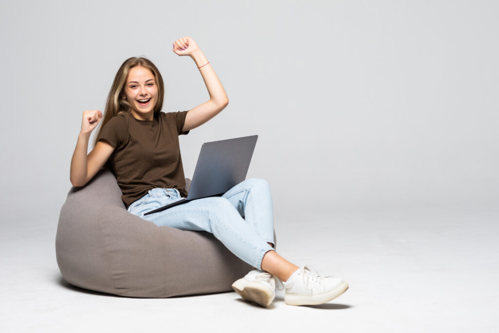 Young woman sitting on pufff with laptop with win gesture on white background